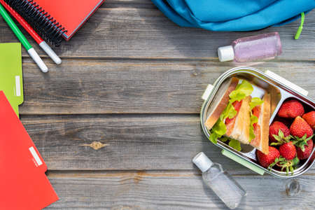 Lunchbox with sandwiches. Box of fresh strawberries, bottles of sanitizer and blue backpack with notebooks and pens on wooden background.Back to school.Lunch with safety precautions after coronavirusの写真素材