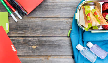 Lunchbox with sandwiches. Box of fresh strawberries, bottles of sanitizer and blue backpack with notebooks and pens on wooden background.Back to school.Lunch with safety precautions after coronavirusの写真素材