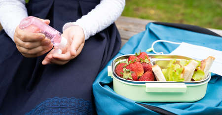 School girl in blue dress pours sanitizer on hands during lunch break on bench in park. Face mask, lunchbox with sandwiches, strawberries. Box on blue backpack. Safety precautions after coronavirusの写真素材