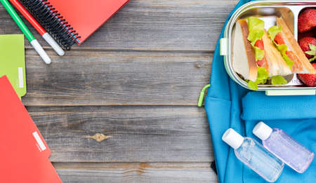 Lunchbox with sandwiches. Box of fresh strawberries, bottles of sanitizer and blue backpack with notebooks and pens on wooden background.Back to school.Lunch with safety precautions after coronavirusの写真素材