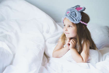 Cute little girl wake up in bed in the morning wearing gray bunny sleep mask. Happy adorable kid...