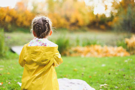 Little girl in yellow raincoat, with pigtails sits on plaid blanket on autumn picnic, looks into...