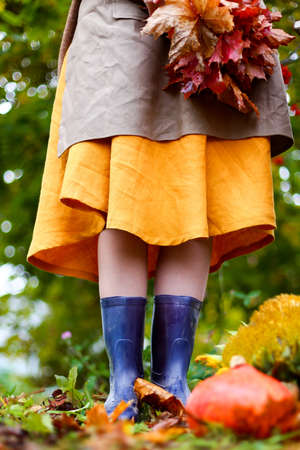 Young woman in yellow dress, cardigan, linen apron, blue rubber boots with bouquet of red maple leaves. Autumn harvest. Cut orange pumpkin, yellow sunflower on grass in garden. Fall style photoshoot.の写真素材