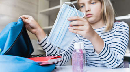 Little girl packing blue backpack in kids's room. Face mask, bottle of sanitizer, stationery, pens, multicolored markers, notebooks. Back to school. Mom's hygiene, safety precautions after coronavirus.の写真素材