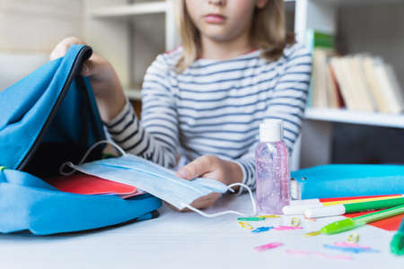 Little girl packing blue backpack in kids's room. Face mask, bottle of sanitizer, stationery, pens, multicolored markers, notebooks. Back to school. Mom's hygiene, safety precautions after coronavirus.の写真素材