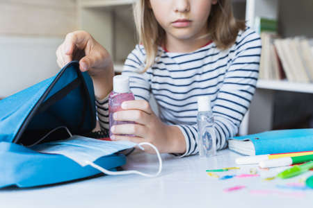 Little girl packing blue backpack in kids's room. Face mask, bottle of sanitizer, stationery, pens, multicolored markers, notebooks. Back to school. Mom's hygiene, safety precautions after coronavirus.の写真素材