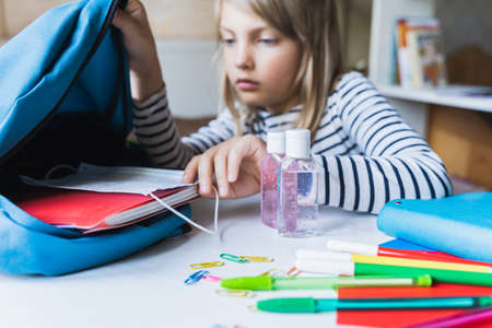 Little girl packing blue backpack in kids's room. Face mask, bottle of sanitizer, stationery, pens, multicolored markers, notebooks. Back to school. Mom's hygiene, safety precautions after coronavirus.の写真素材
