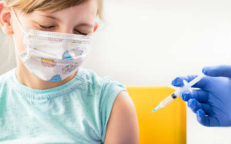 Little girl in face mask in doctor's office is vaccinated. Crying, scared, afraid of syringe with vaccine for covid-19 coronavirus, flu, infectious diseases. Clinical trials of injection for human, child.の写真素材