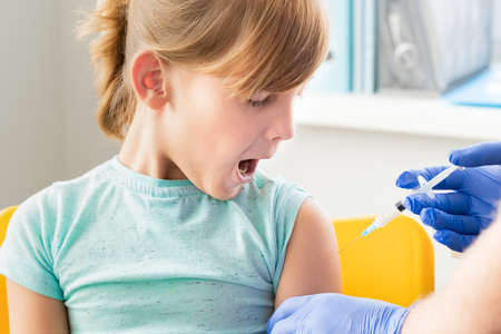 Little girl in face mask in doctor's office is vaccinated. Crying, scared, afraid of syringe with vaccine for covid-19 coronavirus, flu, infectious diseases. Clinical trials of injection for human, child.の写真素材
