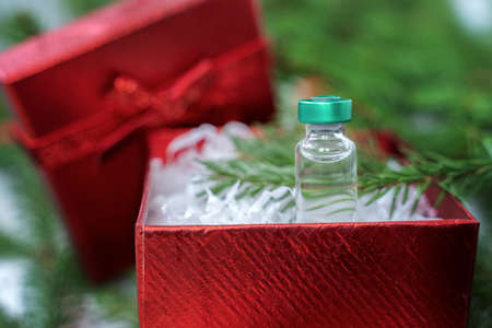 Best gift for Christmas. Vial with new tested vaccine for covid-19 coronavirus, dangerous flu, infection. Small red holiday box with bow. Green branches of new year tree, decorations. White plaid.の写真素材