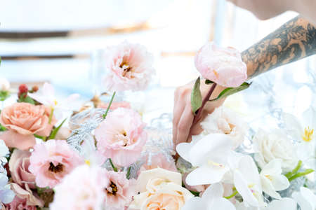 Woman florist on wedding makes flower composition bride and groom from roses, tulips, peonies, orchids on table. Decorators working at event: birthday, anniversary, party. Hand with floral tattoo.のeditorial素材