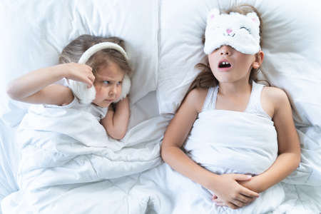 Little girls sleep in bed. One is snoring hard, sister is plugging ears with fur headphones. Early morning wake up, rise to kindergarten, school. Bedtime, sweet dreams.Kids correct daily routine for child.の写真素材