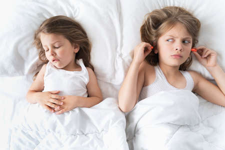 Little girls sleep in bed. One is snoring hard, sister is plugging her ears with earplugs. Early morning wake up, rise to kindergarten, school. Bedtime, sweet dreams.Kids correct daily routine for child.の写真素材