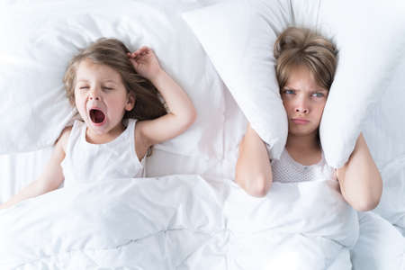 Little girls sleep in bed. One is snoring hard, sister is plugging her ears with pillow. Early morning wake up, rise to kindergarten, school. Bedtime, sweet dreams.Kids correct daily routine for child.の写真素材