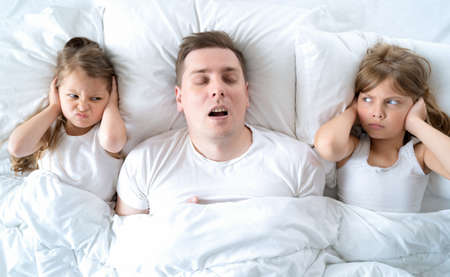 Kids, dad sleep together in bed. Father is snoring hard, girls, daughters are plugging ears with hands. Disorders, joint sleeping. Insomnia, night bedtime. Wake up, rise to kindergarten, school, work.の写真素材