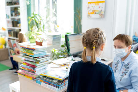 Russia Moscow 25.09.20 Little girl talks to librarian in face mask, asks, loos for book with fairy...