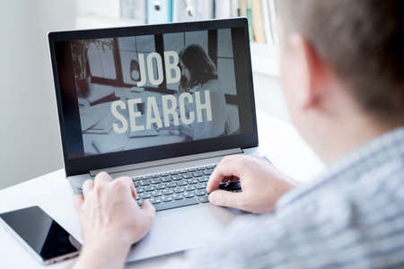 Young man is typing on laptop keyboard, looking for job, telework at home sitting at table. Hiring remote employees, freelancers, specialists for work in business companies in crisis due to coronavirus.の写真素材