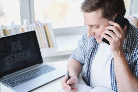 Remote job search. Smiling man speaks on mobile phone, appoints interview for vacancy, writes...