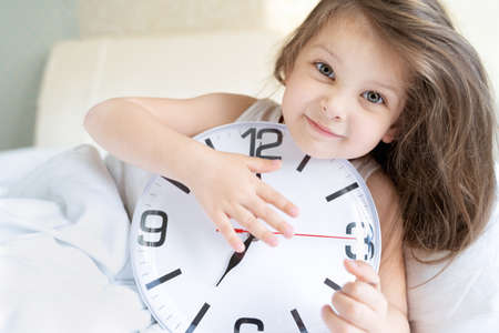 Little sleeping girl with big, huge clock in hands. Early morning wake up before kindergaten, school. White pillow, blanket in bed. Sleepy child. Sweet dreams. Bedtime. Correct mode of children's rest.の写真素材