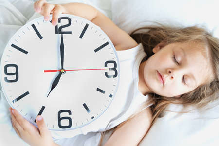 Little sleeping girl with big, huge clock in hands. Early morning wake up before kindergaten, school. White pillow, blanket in bed. Sleepy child. Sweet dreams. Bedtime. Correct mode of children's rest.の写真素材