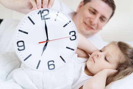 Dad wakes up little sleeping in bed girl, daughter. Big alarm, clock. Early morning before kindergaten, school. White pillow, blanket in bed. Sleepy child.Displeased, sad, unhappy.Bedtime.Daily routine.の写真素材