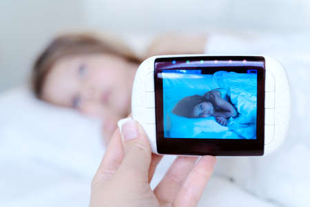 Mom looks at the camera for a little sleeping girl, daughter through the radio, video baby monitor. Protection, care for the safety of the child. Remote control of the soap. Modern technologies.の写真素材