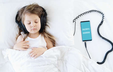 Little girl listening to children's audiobook in mobile phone in headphones before sleeping in bed. Education, child development. E-book with fairy tales, lullaby for the night. Modern technology.の写真素材