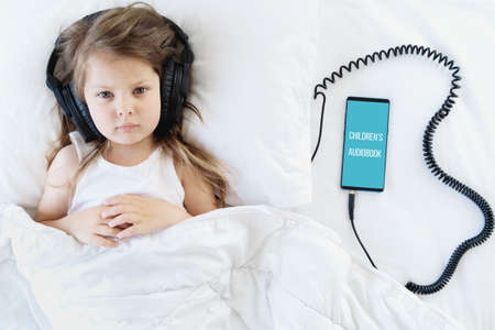 Little girl listening to children's audiobook in mobile phone in headphones before sleeping in bed. Education, child development. E-book with fairy tales, lullaby for the night. Modern technology.の写真素材