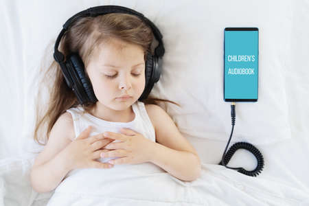 Little girl listening to children's audiobook in mobile phone in headphones before sleeping in bed. Education, child development. E-book with fairy tales, lullaby for the night. Modern technology.の写真素材