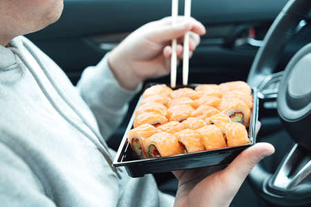 Man is eating sushi, Philadelphia rolls with fish, rice, salmon, cream cheese, avocado in car. Fast, safe, takeaway food on the road to coronavirus pandemic. Delicious japanese snacks with chopsticks.の写真素材