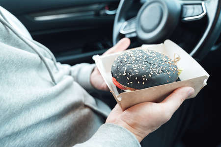Man is eating black burger with meat, fish, tomato, salad, sause in car. Fast, safe, takeaway food...