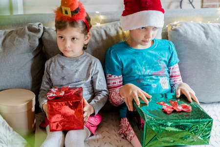 Little cute girls, kids in red Christmas santa claus, elf hat with gifts in hands from family, parents.Congratulations, greetings.New Years celebration in country wooden house on coronavirus isolation.の写真素材