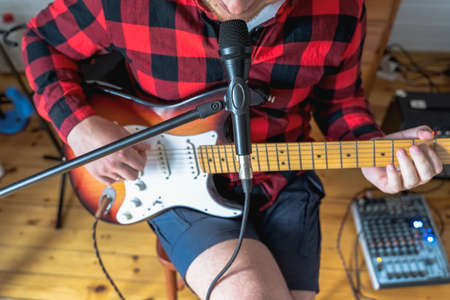 Young handsome man, musician, singer studying, practicing to play electric guitar at home, singing songs into microphone.Hobby, passion.Guy in red plaid shirt sits on chair.DJ console, speakers, equipment.の写真素材