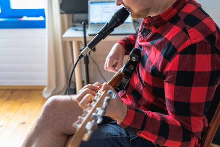 Young handsome man, musician, singer studying, practicing to play electric guitar at home, singing songs into microphone.Hobby, passion.Guy in red plaid shirt sits on chair.DJ console, speakers, equipment.の写真素材