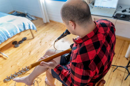 Young handsome man, musician, singer studying, practicing to play electric guitar at home, singing songs into microphone.Hobby, passion.Guy in red plaid shirt sits on chair.DJ console, speakers, equipment.の写真素材