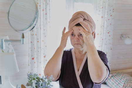 Aged, elderly beutiful woman in bathrobe and towel on head applies cosmetics, cream, does facial massage. Self care.Treatment for aging skin.Women's cozy wooden bedroom, chest of drawers, mirror, boudoir.の写真素材