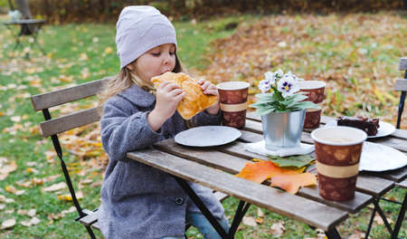 Little cute girls, sisters in coats seat, eat buns, drink coffee in cafe in autumn park. Cozy wooden table, chairs. Drinks, food to go, take away.Maple leaves, foliage.Outdoor family activity in nature.の写真素材