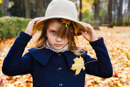 Little cute stylish long hair girl in autumn coat holds hat with multi-colored yellow red maple leaves, foliage in hands. Walking, playing funny games in park. Outdoor family activity in nature.の写真素材