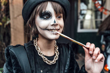 Beautiful scary little girl celebrating halloween, chewing gummy worm. Terrifying face makeup and...