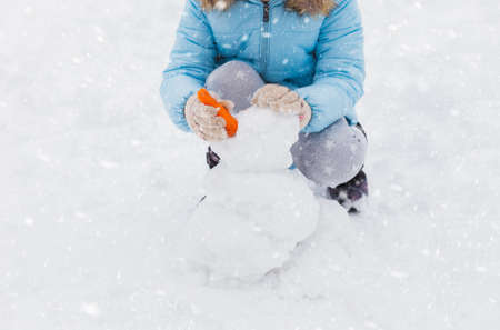 Little girl is making snowman from snowballs with carrot in snowy winter park. Walking, having fun.Stylish jacket with fur, pants with snowflakes, mittens.Family picnic in cold weather.Outdoor activity.の写真素材