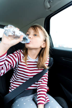 Little caucasian girl is driving in car, dreanking pure water from plastic kids bottle. Traveling on road in safe baby seats with child belts. Fun family trip, activity with parents.の写真素材