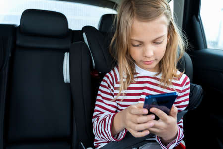Little cute caucasian girl is driving in car. Kid writing sms, chatting, playing video games in mobile phone. Traveling on road in safe baby seat with child belt.Fun family trip, activity with parents.の写真素材