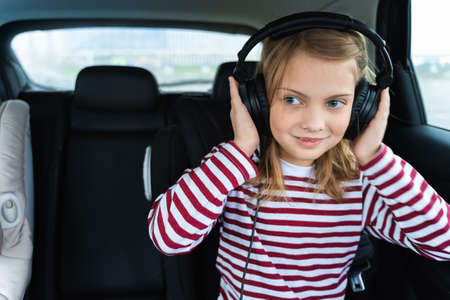 Little cute caucasian girl is driving in car.Kid listening to music with big headphones.Traveling,...