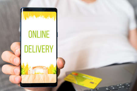 Hand with mobile phone. Online internet shopping, order through mobile application. Laptop. Home delivery of food, groceries from grocery store. Paper bag with pasta on screen. Payment by credit card.の写真素材