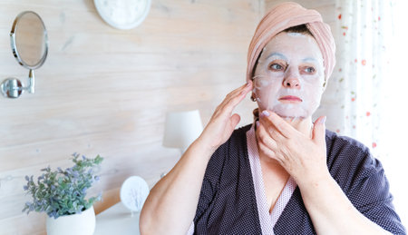 Aged, elderly woman in bathrobe uses cosmetic sheet face mask, applies cosmetics. Self care. Treatment for aging skin. Women's cozy wooden bedroom, chest of drawers, mirror, boudoir.の写真素材