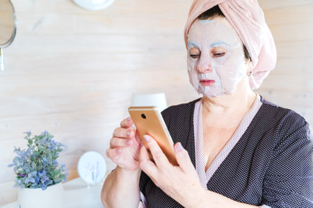 Aged, elderly woman in bathrobe uses cosmetic sheet face mask and speaks by mobile phone. Self care. Anti aging treatment for skin. Women's cozy wooden bedroom, chest of drawers, mirror, boudoir.の写真素材
