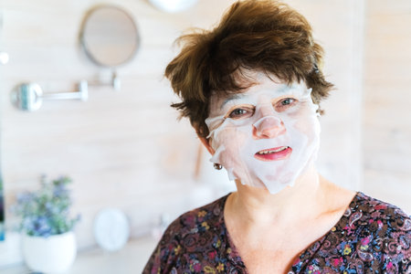 Funny aged, elderly woman with cosmetic sheet face mask. Application of cosmetics. Self care. Treatment for aging skin. Women's cozy wooden bedroom, chest of drawers, mirror, boudoir.の写真素材
