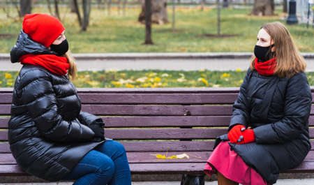 Two young women, friends in face masks speak to each other on safe social distance sitting on bench...