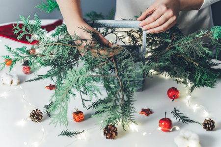 Florist woman makes Christmas composition for table decoration from thuja, spruce branches in wooden box. Using of floral foam, cones, snow apples, balls, cotton flowers,garland. New Year celebration.の写真素材