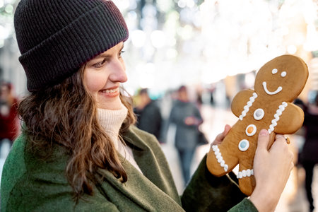Young beautiful girl, woman with huge funny gingerbread man. Christmas shopping. Walking on market street in big city decorated with garlands, lights.Stylish look, wool coat, hat.New year celebration.の写真素材